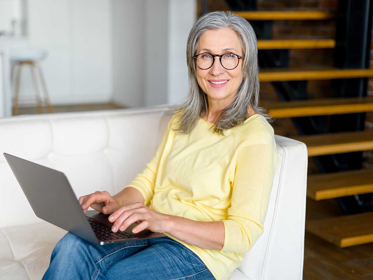 Positive charming mature gray-haired woman in glasses sitting on the sofa, typing on laptop, doing online shopping, working on a project, watching webinar. Freelancer enjoying remote work from home